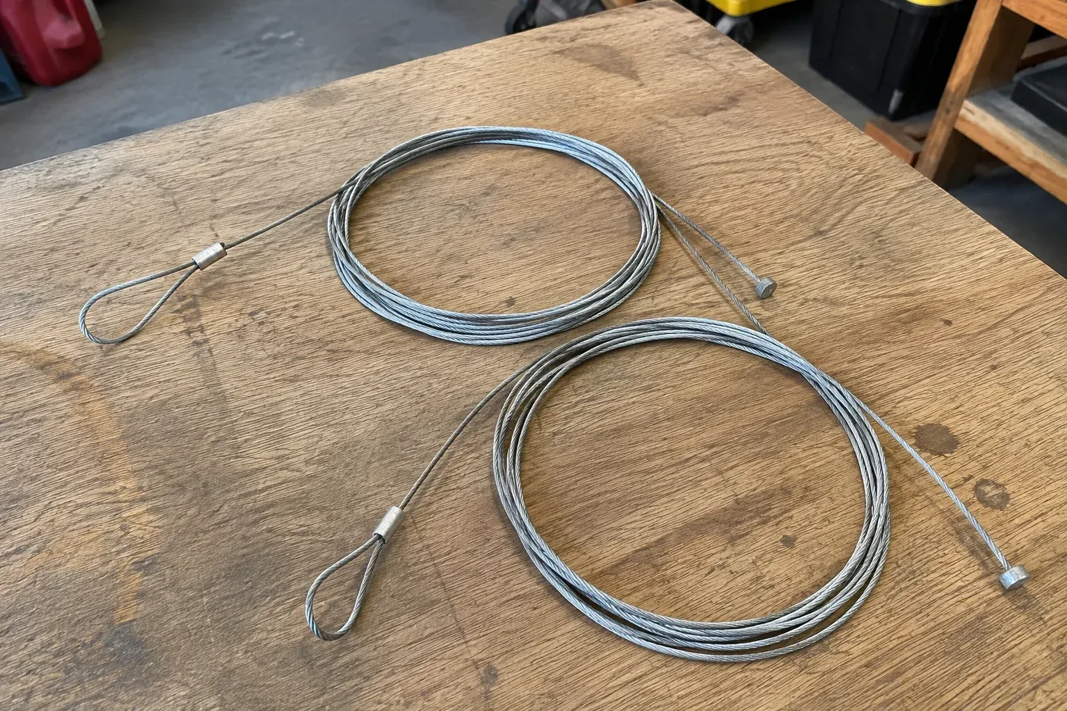 Two coiled steel cables with loop and stop ends on a workbench. — Garage Door Cable Replacement, Essa
