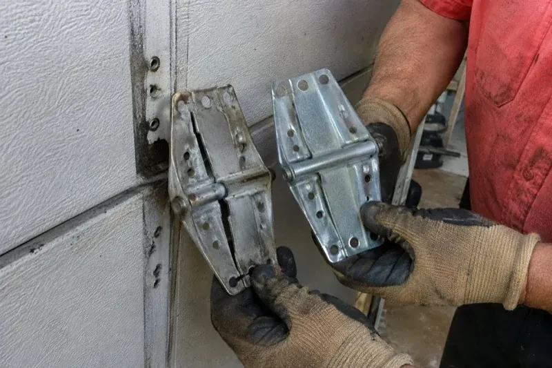 Gloved hands holding two metal garage-door hinges side by side, one discolored with a visible crack and the other bright silver. — Garage Door Hinge Replacement, Essa