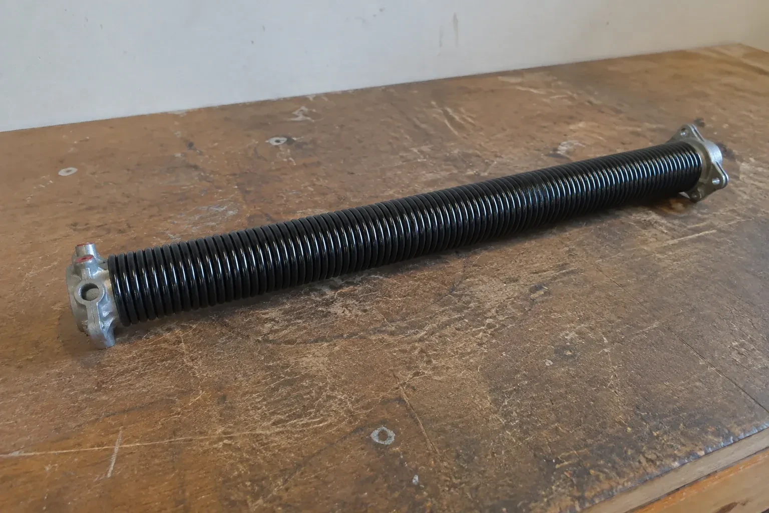 Detached black coil spring with metal end fittings on a workbench. — Torsion Spring Replacement, Essa